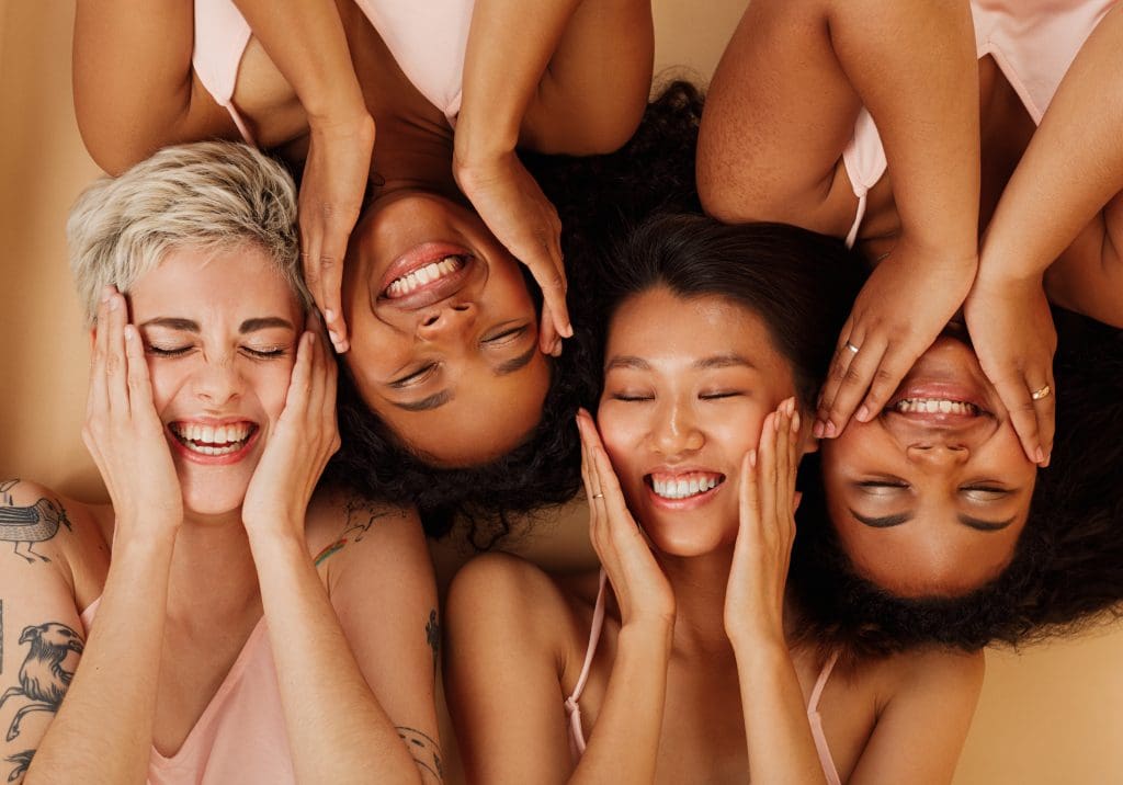 4 women with closed eyes, smiling and rubbing their cheeks while laying next to each other, as part of National Self-Care Day