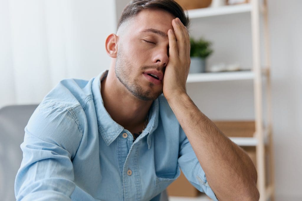 a tired, frustrated man suffering from chronic fatigue syndrome sits with his hand on his face