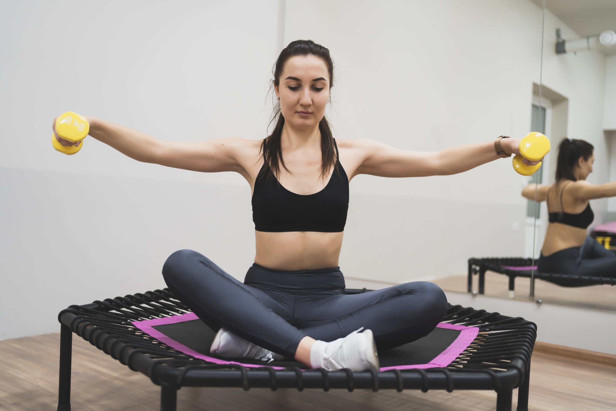 a woman sitting on a mini trampoline while holding weights utilizing rebounder therapy for weight loss