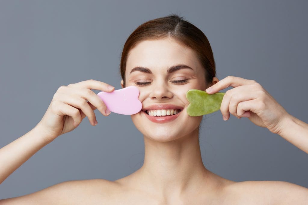 a smiling woman using a gua sha stone on each side of her face agaisnt a gray background to show the gua sha results timeline